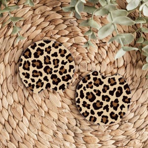 May include: Two brown and black leopard print car coasters. The coasters are round and have a cut-out shape for a cup holder.