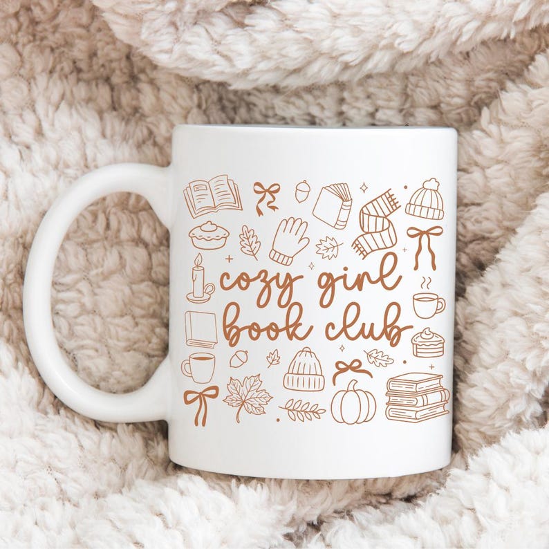 May include: White ceramic mug with a brown illustration of fall-themed icons and the words "cozy girl book club." The design includes books, leaves, a pumpkin, and a hat. The mug has a curved handle.