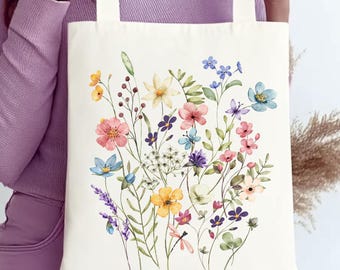 May include: Cream-colored tote bag featuring a watercolor floral design. The artwork showcases a vibrant array of flowers in shades of pink, yellow, blue, and purple, with green foliage. The bag has cream-colored handles.