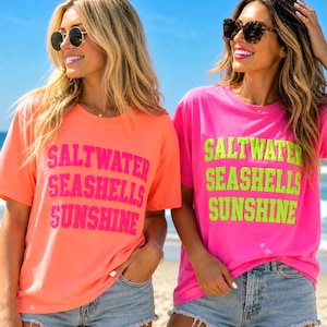 以下が含まれることがあります： サングラスをかけた2人の女性が、「SALTWATER, SEASHELLS, SUNSHINE」とプリントされたTシャツを着ています。1枚はピンクの文字が入ったコーラル、もう1枚はネオングリーンの文字が入ったホットピンクです。どちらもデニムショーツを着用しています。