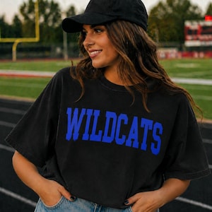May include: Black t-shirt with the word "WILDCATS" in bold blue letters. The shirt is worn with a black baseball cap. The background shows a football field.