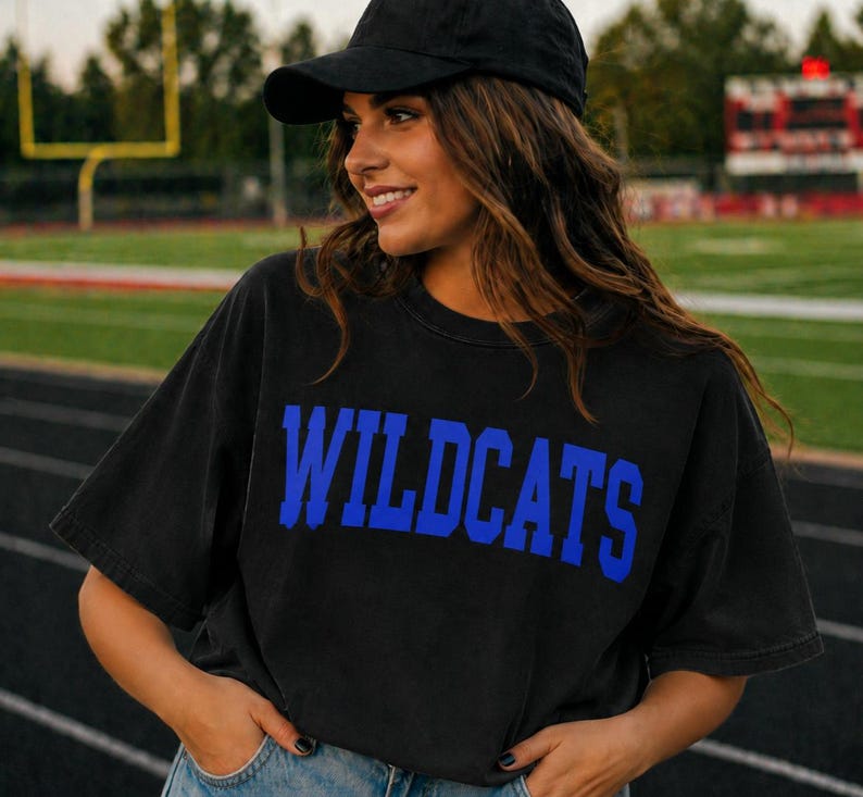May include: Black t-shirt with the word "WILDCATS" in bold blue letters. The shirt is worn with a black baseball cap. The background shows a football field.