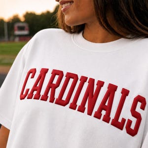 May include: White sweatshirt with the word "CARDINALS" in bold, red, block letters across the chest. The sweatshirt has a crew neck and short sleeves. The background is a blurred outdoor scene.