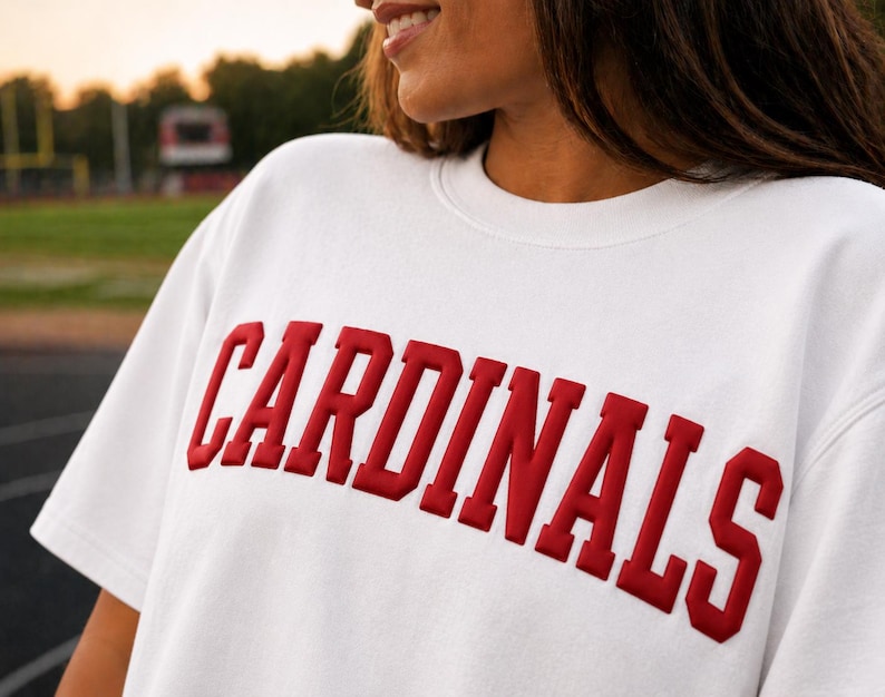May include: White sweatshirt with the word "CARDINALS" in bold, red, block letters across the chest. The sweatshirt has a crew neck and short sleeves. The background is a blurred outdoor scene.
