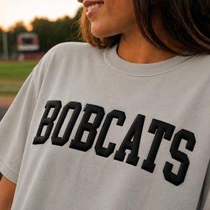 May include: A light gray t-shirt with the word "BOBCATS" in bold, black, block letters across the chest. The shirt has short sleeves and a crew neck. The background shows a blurred outdoor setting.