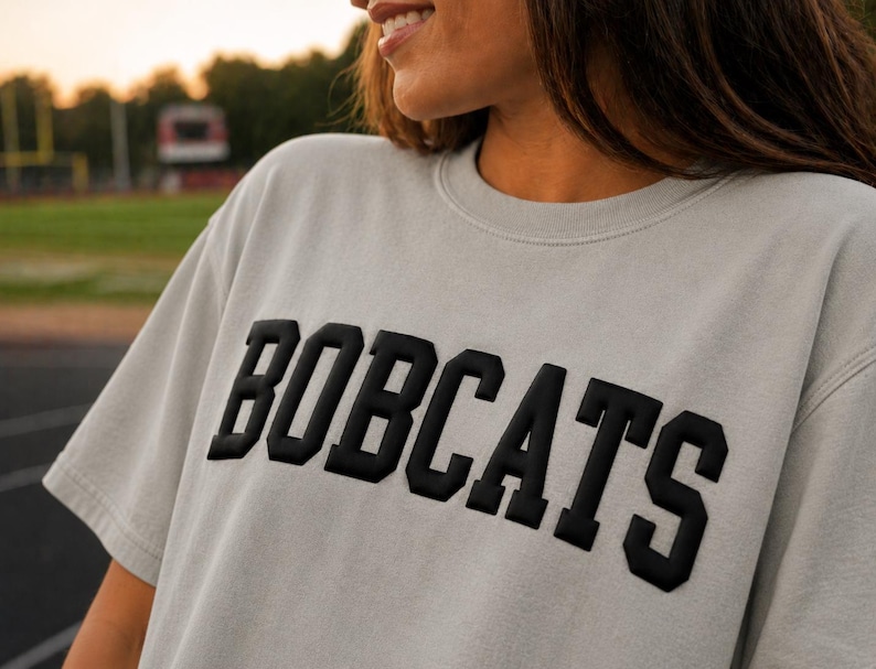 May include: A light gray t-shirt with the word "BOBCATS" in bold, black, block letters across the chest. The shirt has short sleeves and a crew neck. The background shows a blurred outdoor setting.