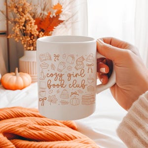 May include: White ceramic mug with the text "cozy girl book club" and autumn-themed illustrations. The mug features drawings of books, mittens, hats, and pumpkins. The mug is held by a person.