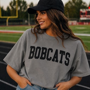 May include: Gray t-shirt with the word "BOBCATS" in black block letters. The shirt is worn with a black baseball cap. The image is taken outdoors on a track field.