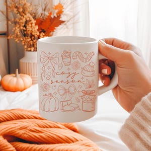 May include: White ceramic mug with a handle featuring a "cozy season" design in orange. The design includes bows, a coffee cup, a scarf, a hat, a pumpkin, and other fall-themed illustrations. The mug is held by a person.