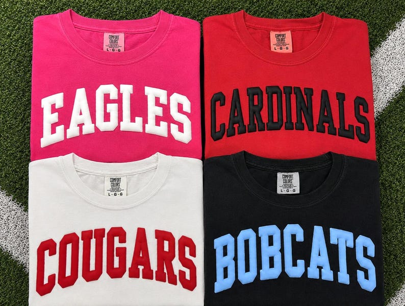 May include: Four t-shirts in pink, red, white, and black, each with a different team name in bold, block letters: Eagles, Cardinals, Cougars, and Bobcats. The shirts are laid out on a green surface with a white stripe.