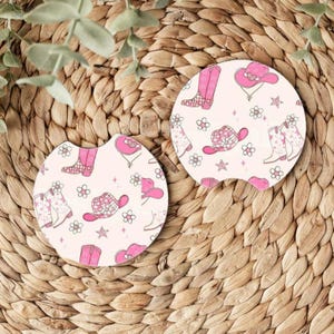 May include: Two pink and white car coasters with a pattern of cowboy boots and hats. The coasters are round and have a white background with pink accents.