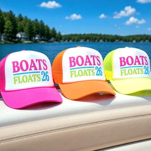 May include: Three trucker hats with white mesh backs and colored brims. Each hat has the words "BOATS FLOATS 26" in pink, blue, and green. The hats are pink, orange, and yellow, and are sitting on a boat.