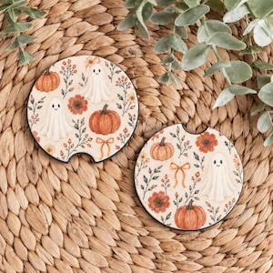 May include: Two circular car coasters featuring a cream-colored background with a repeating pattern of orange pumpkins, white ghosts, orange flowers, green foliage, and small orange bows. Each coaster has a small semi-circular notch at the top edge.