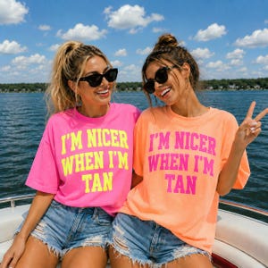 Pode incluir: Duas mulheres sorrindo usam óculos de sol e camisetas com o texto "I'M NICER WHEN I'M TAN". Uma camisa é rosa brilhante com texto amarelo, a outra é laranja com texto rosa. Em um barco.