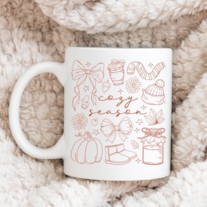 May include: White ceramic mug with a brown illustration of fall-themed drawings, including bows, a coffee cup, a scarf, and a pumpkin. The words "cozy season" are written in a script font.