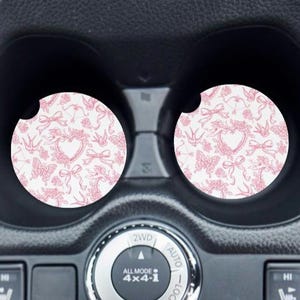 May include: Two pink and white car coasters with a floral and heart design. The coasters are in the cup holders of a car.