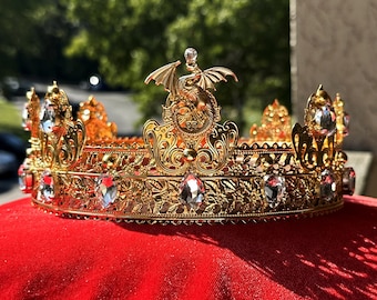 May include: A gold crown with a dragon design on top and clear crystal accents. The crown is on a red velvet surface.