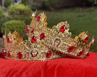 May include: Two ornate gold crowns with red gemstones and religious icons. The crowns are placed on a red velvet surface, with a blurred green background. The crowns have intricate filigree designs and are likely used for religious ceremonies.