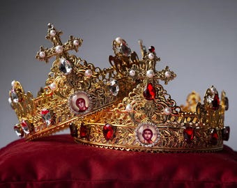 Puede incluir: Dos coronas doradas ornamentadas con filigrana metálica intrincada, rematadas con cruces y pequeñas perlas blancas. Cada corona está adornada con gemas facetadas transparentes y de color rojo intenso, e iconos circulares con figuras.