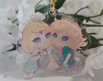 May include: A clear acrylic keychain featuring two anime characters, a blonde woman with a green dress and a blonde man with a blue and green outfit. The characters are holding hands and standing in front of a pink and white triangle symbol. The keychain has a gold chain.