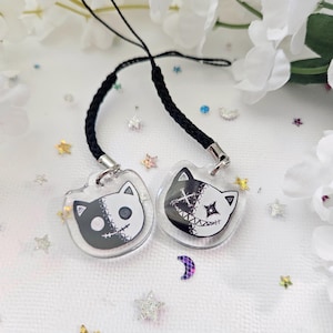May include: Two clear acrylic cat charms with black and white stitched designs. Each charm has a black braided cord and silver-tone metal clasp. The cat faces are split, with one side black and the other white, featuring stitched details.