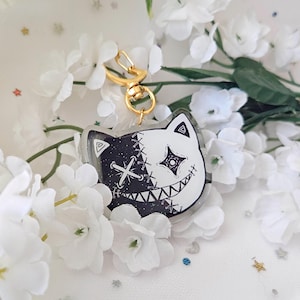 May include: A black and white cat-shaped keychain with a stitched design and a gold-colored clasp. The cat's face is split, with one side black and the other white, featuring a stitched cross and a star-shaped eye. The keychain is surrounded by white flowers.