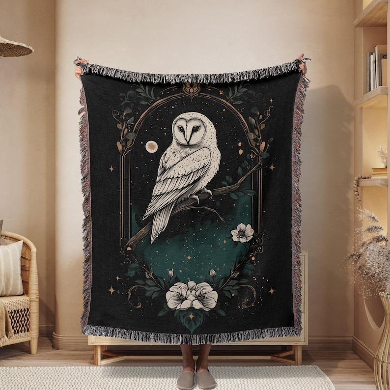 May include: A black woven throw blanket with a white owl perched on a branch with flowers. The owl is surrounded by a gold frame with a dark green background and stars.