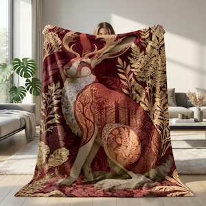 Könnte beinhalten: Eine große rechteckige Wohndecke mit tiefrotem Hintergrund und einer zentralen Hasenillustration. Der Hase ist mit komplexen goldfarbenen Baum- und Blattmustern verziert und von goldenem botanischem Blattwerk sowie Farnen umgeben.