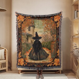 May include: A woven tapestry throw blanket features a witch walking through a forest path with autumn leaves. The witch is wearing a black dress and a pointed hat. The background is a house with a gate and trees with orange and yellow leaves.