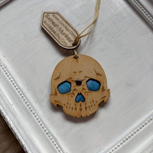 May include: Wooden skull decoration with blue eye inserts and a gold hanging string. The ornament features a detailed laser-cut design and a small wooden tag with text. The tag reads: Bunneah Munkeah, and BunneahMunkeah.Etsy.Com.