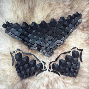 May include: A set of black, geometric armour pieces, comprising a chest plate and two arm guards, are shown against a textured, light-coloured backdrop. The armour is made from small, dark, square-shaped elements.