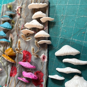 以下が含まれることがあります： 花柄の刺繍が施されたグレーの生地に、白、青、ピンク、ゴールドの色合いのキノコ型の装飾が施されています。キノコは装飾的なパターンで配置されています。