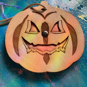 May include: Wooden pumpkin decoration with carved face details, including triangular eyes and a jagged mouth. The pumpkin has a natural wood grain appearance with a metal button nose and a brown cord for hanging. The background is a colorful, glittery surface.