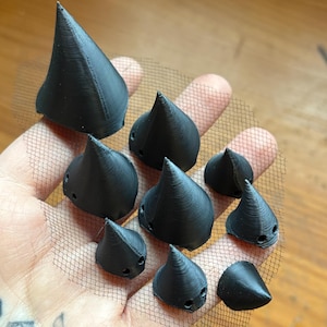 以下が含まれることがあります： 黒色の円錐形オブジェクトのコレクション。表面はテクスチャ加工されており、おそらく3Dプリントされています。黒いメッシュの上に配置され、サイズに若干のばらつきがあり、小さな穴があります。背景は木材です。