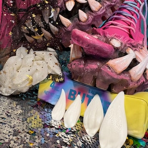 May include: A pair of bright pink lace-up boots featuring sculpted monster teeth along the soles and tongues. The boots are displayed with a black star-patterned mesh bag, a white star-patterned fabric, and four smooth white fangs resting on a yellow box.