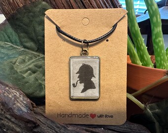 May include: A rectangular pendant necklace featuring a black silhouette of a person with a pipe and hat, likely Sherlock Holmes. The pendant hangs from a black cord and is attached to a brown card with the words "Handmade with love".
