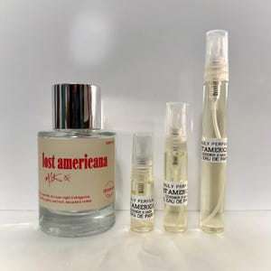 May include: A perfume bottle and three sample vials are displayed against a white background. The large bottle has a silver cap and a label with the text "lost americana" in red. The sample vials have labels with the text "Truly Perfume America".