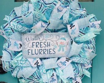 May include: A winter wreath featuring a sign with the text "Winter Fresh Flurries" and a snowman design. The wreath is crafted from light blue and white ribbons adorned with snowman and snowflake motifs. It is displayed on a teal door.