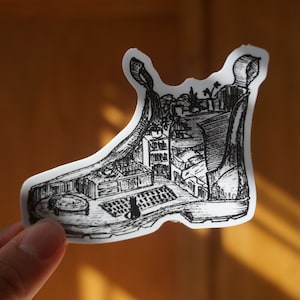May include: A black and white illustration of a boot with a detailed interior scene. The interior includes furniture, plants, and a cat. The artwork is a sticker, held by a hand, with a warm, natural background.