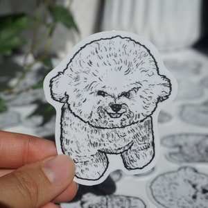 Op de afbeelding: Een zwart-wit sticker met een cartoon illustratie van een pluizige hond. De hond heeft een vriendelijke uitdrukking en staat rechtop. De sticker wordt door een hand vastgehouden, met andere soortgelijke stickers zichtbaar op de achtergrond.