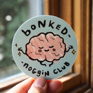 Peut inclure: Un autocollant rond avec le texte "BONKED - NOGGIN CLUB" encerclant un cerveau de dessin animé. Le cerveau est rose avec un visage souriant et des bras. Le fond est bleu clair avec des étoiles.