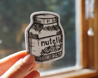 May include: A black and white sticker of a Nutella jar. The sticker is hand-drawn with a sketched style, featuring the word "nutella" in a handwritten font. The jar design includes details like the lid and a depiction of hazelnuts.