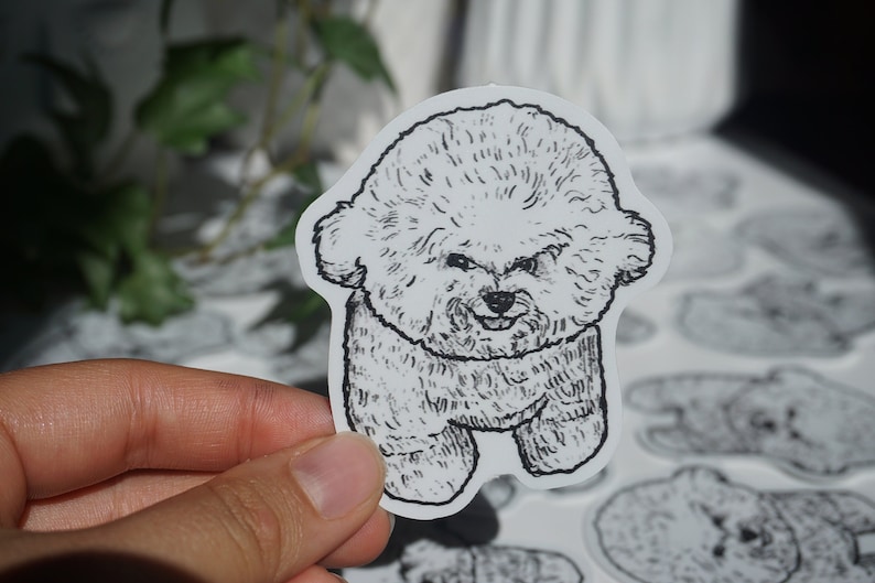 Op de afbeelding: Een zwart-wit sticker met een cartoon illustratie van een pluizige hond. De hond heeft een vriendelijke uitdrukking en staat rechtop. De sticker wordt door een hand vastgehouden, met andere soortgelijke stickers zichtbaar op de achtergrond.