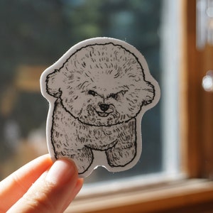 Op de afbeelding: Een gestanste sticker met een gedetailleerde zwart-wit illustratie van een Bichon Frise hond. De sticker heeft een witte rand en toont de hond in een staande pose met een glimlachende uitdrukking. De sticker wordt voor een raam gehouden.