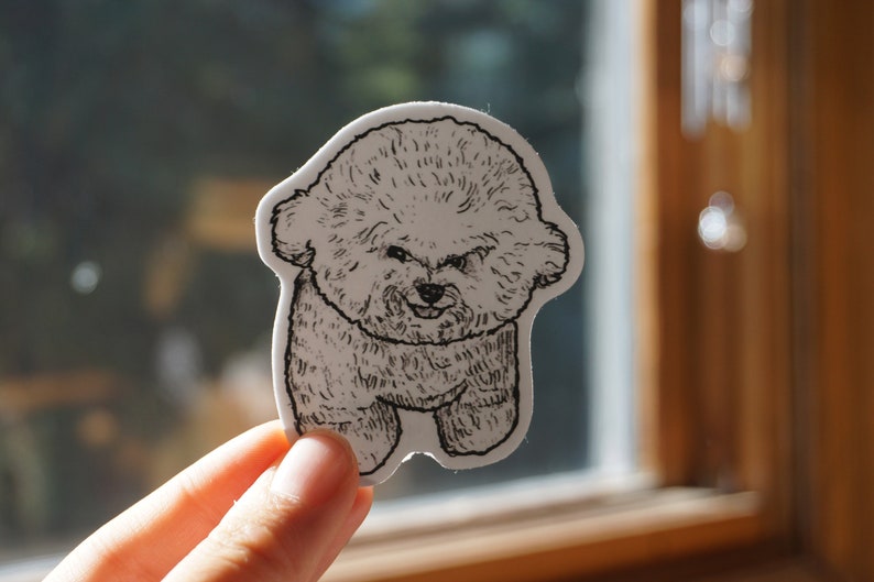 Op de afbeelding: Een gestanste sticker met een gedetailleerde zwart-wit illustratie van een Bichon Frise hond. De sticker heeft een witte rand en toont de hond in een staande pose met een glimlachende uitdrukking. De sticker wordt voor een raam gehouden.