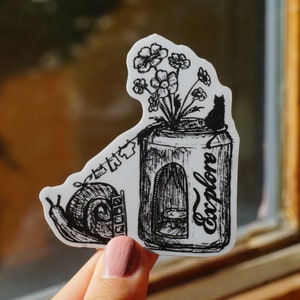 May include: Black and white sticker featuring a whimsical illustration. A can with the word "explore" is filled with flowers and a small black cat. A snail and clothesline are also depicted. The sticker is held by a person.