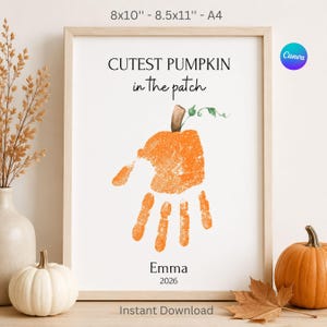 May include: A framed wall art print featuring the text "CUTEST PUMPKIN in the patch" above an orange handprint shaped like a pumpkin with a green stem. Below the handprint is the name "Emma" and the year "2026". Available in 8x10 and 8.5x11 inches.