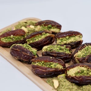 May include: A wooden serving tray with dates filled with pistachio paste. The dates are arranged in a row on the tray, with pistachio powder sprinkled around them.