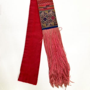 K&ouml;nnte beinhalten: Ein rotes Stoffband mit einem rosa Fransenbesatz und einem bunten bestickten Abschnitt. Die Stickerei zeigt geometrische Muster und florale Motive in Blaut&ouml;nen, Rosa, Gelb und Gr&uuml;n.