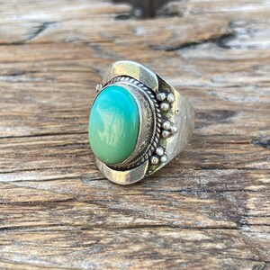 May include: A silver ring featuring a light turquoise oval gemstone. The ring has a detailed setting with small beaded accents and a decorative border. The ring is set against a wooden surface.